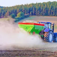 Traktor z rozsiewaczem wapna wykonuje odkwaszanie gleby na polu, tworząc chmurę pyłu.